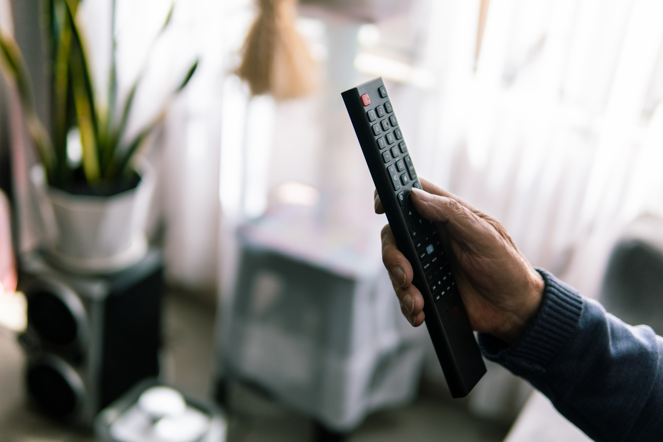 A close-up shot of an elderly man's hand holding a TV remote control with soft focus on the background